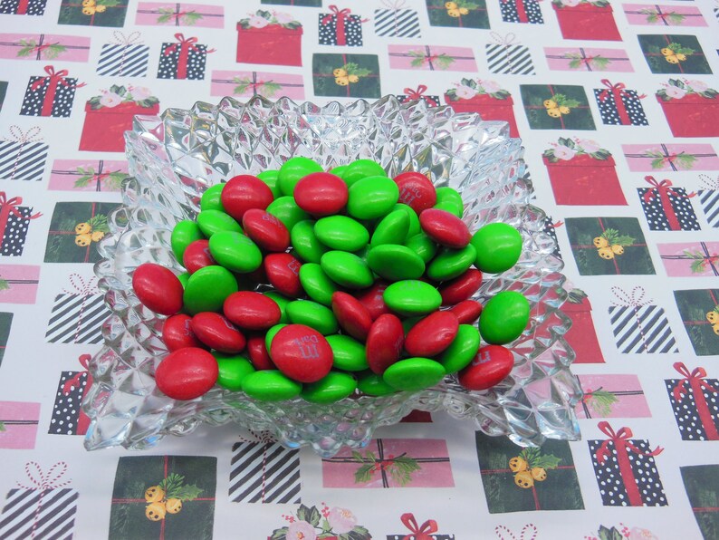 Puede incluir: Un taz&oacute;n de vidrio transparente lleno de caramelos M&M's rojos y verdes. El taz&oacute;n est&aacute; sobre un fondo blanco con un patr&oacute;n repetitivo de cajas de regalo rojas y verdes.