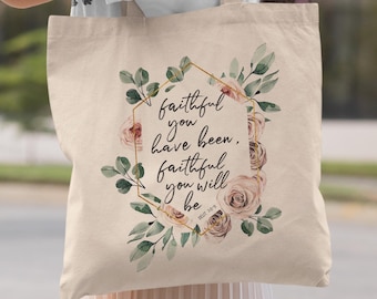 May include: A beige canvas tote bag featuring a gold hexagonal frame surrounded by watercolor-style pink roses and green leaves. The text inside the frame reads, "faithful you have been, faithful you will be" followed by the small citation "DEUT 7:9".