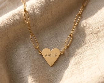 May include: A gold-toned necklace featuring a heart-shaped pendant engraved with the word ABIDE. The pendant is attached to a chain composed of elongated, oval-shaped links. The metal has a smooth, polished finish that reflects light.