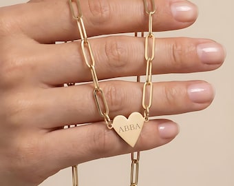 May include: A gold-toned necklace featuring a series of elongated oval links connected to a central heart-shaped pendant. The pendant is engraved with the text ABBA in a clean, serif font, creating a balanced and uniform appearance across the chain.