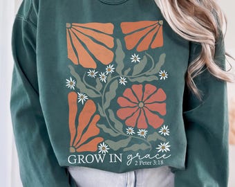 May include: A dark green crewneck sweatshirt featuring a graphic print of orange and muted green abstract flowers and leaves. The text "GROW IN grace 2 Peter 3:18" is printed in white at the bottom of the floral design on the front of the garment.