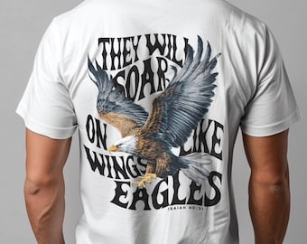 May include: White t-shirt featuring a detailed illustration of an eagle with outstretched wings. The design includes the text "THEY WILL SOAR ON WINGS LIKE EAGLES" and "ISAIAH 40:31" in black. The eagle has brown, white, and blue feathers.