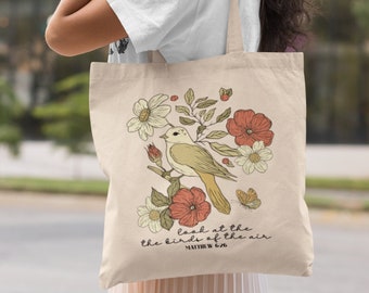 May include: A beige canvas tote bag featuring a printed illustration of a bird perched on a branch surrounded by flowers and a butterfly. The text reads, "look at the birds of the air MATTHEW 6:26" in a black cursive and block font style.