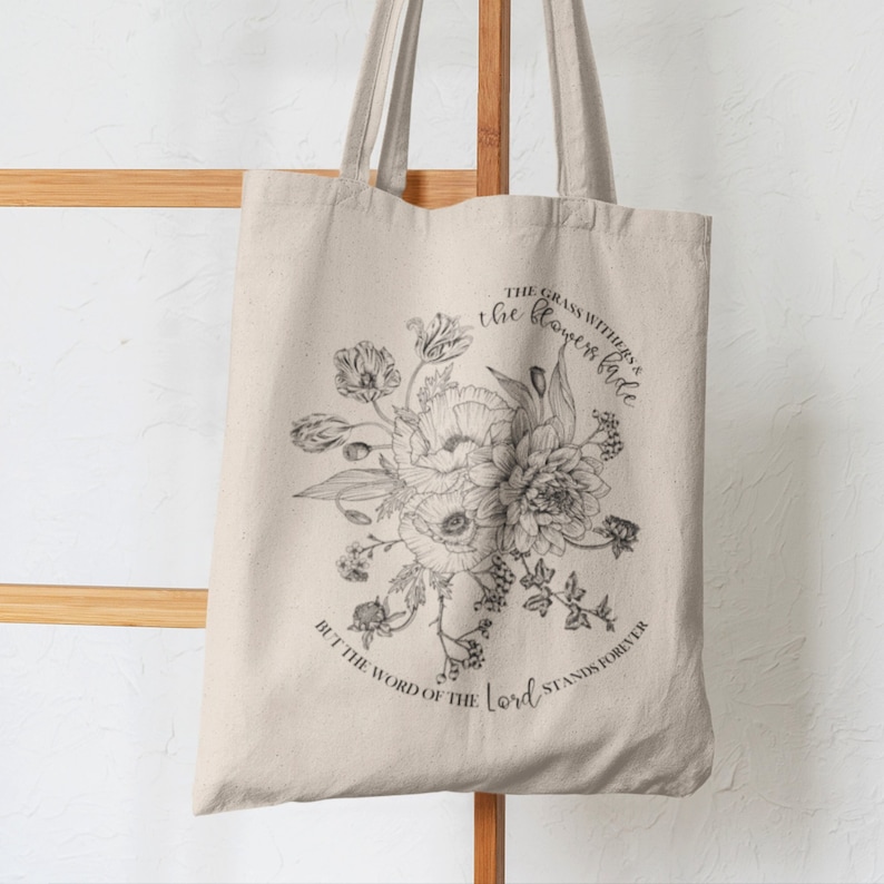 Pode incluir: Uma bolsa tote de lona bege com estampa floral preta e o texto "THE GRASS WITHERS & the flowers fade BUT THE WORD OF THE Lord STANDS FOREVER".