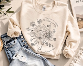May include: A cream-colored crewneck sweatshirt featuring a black line-art floral bouquet illustration. The text reads, The grass withers & the flowers fade, but the word of the Lord stands forever. The sweatshirt is paired with distressed blue denim jeans.