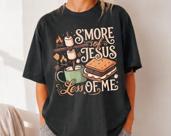 May include: A black short-sleeve t-shirt featuring a graphic print of a s'more, a mug, and a campfire. The text reads S'MORE of JESUS LESS OF ME in a decorative font. The design includes illustrations of marshmallows, chocolate, and graham crackers.