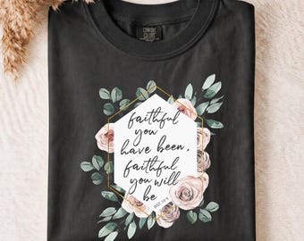 May include: A black crewneck t-shirt featuring a white hexagonal graphic with the text "faithful you have been, faithful you will be" and "DEUT 7:9". The design is surrounded by a gold geometric border and illustrations of pink roses and green leaves.