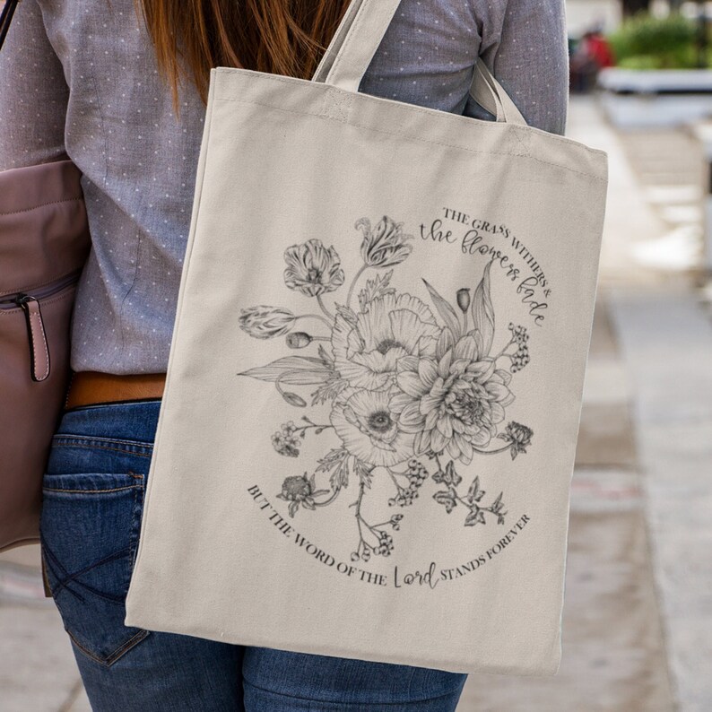 Pode incluir: Uma bolsa tote de lona branca com um design floral preto e branco e o texto "The grass withers & the flowers fade, but the word of the Lord stands forever".