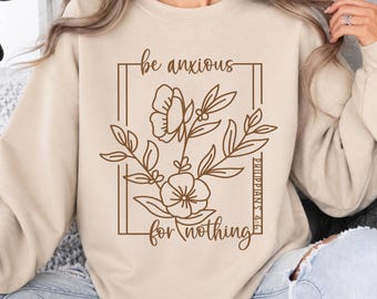 May include: A beige crewneck sweatshirt featuring a brown line art graphic of flowers enclosed in a rectangular frame. The text reads be anxious for nothing and includes the scripture reference Philippians 4:6 in a vertical orientation.