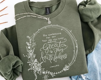 May include: An olive green crewneck sweatshirt featuring a white graphic print of a floral wreath surrounding the text: His compassions fail not. They are new every morning: Great is Thy faithfulness. Lam. 3:22-23. A silver chain necklace rests on top.