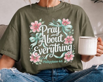 May include: An olive green t-shirt featuring a graphic print with the text "Pray About Everything" and "Philippians 4:6". The design includes pink flowers, green leaves, and light blue ribbons arranged in a decorative square frame.