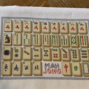 May include: A cross-stitch embroidery of a Mahjong set with the words "Mah Jong" in red and white. The tiles are arranged in rows and columns, with each tile featuring a different symbol or number. The background is a cream-colored fabric.