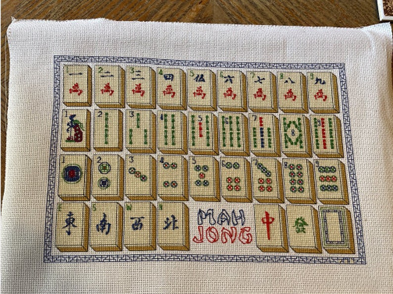 May include: A cross-stitch embroidery of a Mahjong set with the words "Mah Jong" in red and white. The tiles are arranged in rows and columns, with each tile featuring a different symbol or number. The background is a cream-colored fabric.