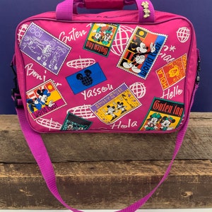 May include: Pink travel bag with a Mickey Mouse theme. The bag features a variety of colorful patches with Mickey Mouse and Minnie Mouse, along with greetings in different languages, including "Guten Tag", "Hola", "Yassou", and "World Tour".