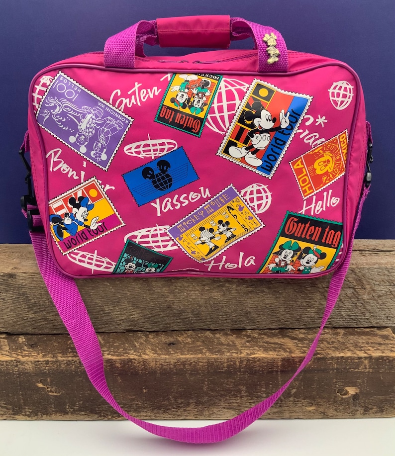 May include: Pink travel bag with a Mickey Mouse theme. The bag features a variety of colorful patches with Mickey Mouse and Minnie Mouse, along with greetings in different languages, including "Guten Tag", "Hola", "Yassou", and "World Tour".