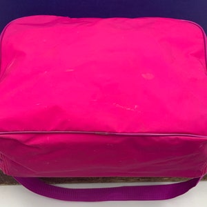 May include: A bright pink, rectangular, zippered bag with a purple strap. The bag has a smooth, shiny finish and is made of a lightweight material.