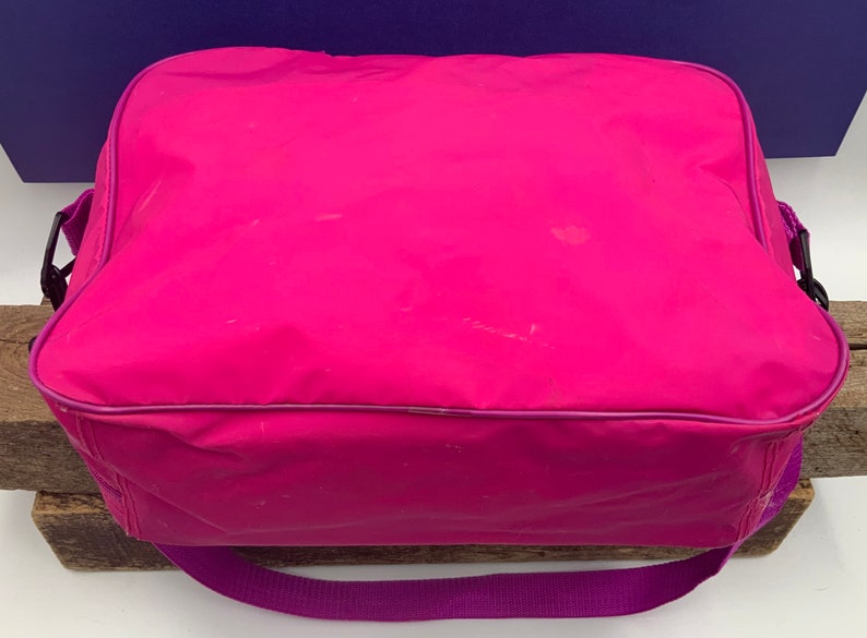 May include: A bright pink, rectangular, zippered bag with a purple strap. The bag has a smooth, shiny finish and is made of a lightweight material.