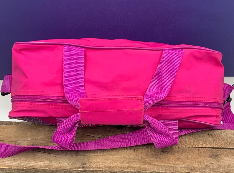 May include: A pink travel bag with a zippered closure and two purple straps. The bag has a small pocket on the front.