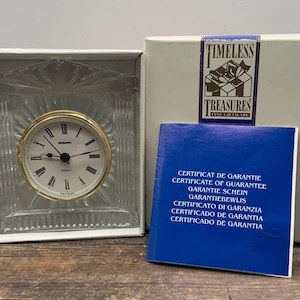 May include: A white box with a crystal clock inside. The clock has a gold face with black hands and Roman numerals. The box has a blue label with the text "TIMELESS TREASURES FINE GIFTW ARE" and a certificate of guarantee in multiple languages.