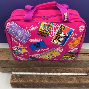 May include: Pink travel bag with a Mickey Mouse theme. The bag has a zipper closure and a top handle. The bag is decorated with colorful stamps featuring Mickey Mouse and the words "Guten Tag", "Yassou", "Hello", and "World Tour".