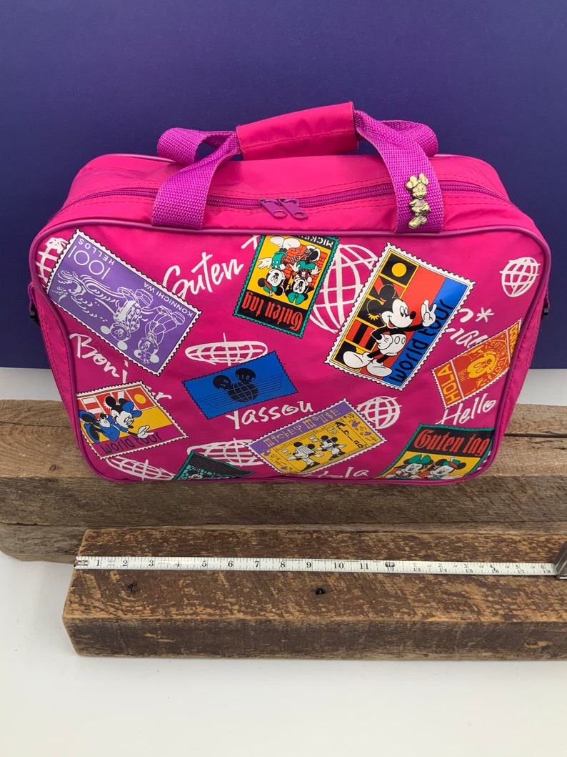 May include: Pink travel bag with a Mickey Mouse theme. The bag has a zipper closure and a top handle. The bag is decorated with colorful stamps featuring Mickey Mouse and the words "Guten Tag", "Yassou", "Hello", and "World Tour".