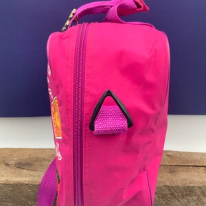 May include: A pink travel bag with a purple strap and a black zipper. The bag has a small black handle on the side and a graphic of a cartoon character on the front.
