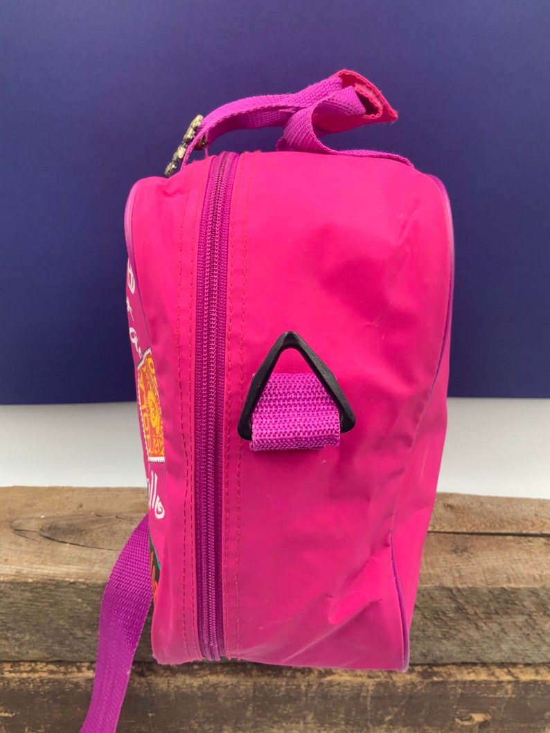 May include: A pink travel bag with a purple strap and a black zipper. The bag has a small black handle on the side and a graphic of a cartoon character on the front.
