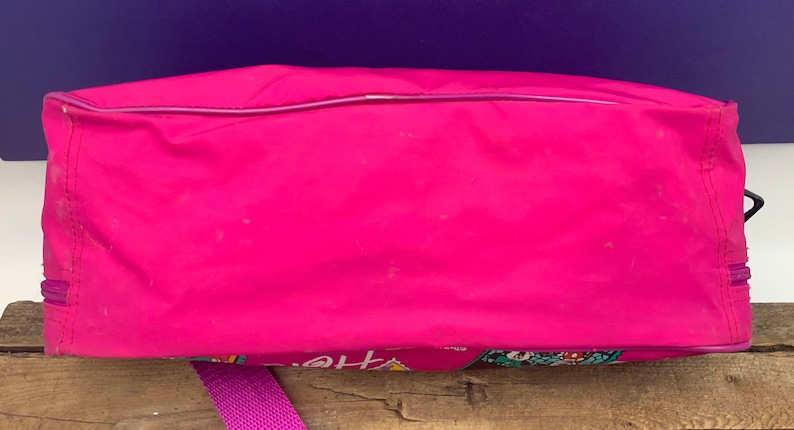 May include: A pink nylon tote bag with a purple zipper and a colorful graphic print on the bottom. The bag has a purple strap.