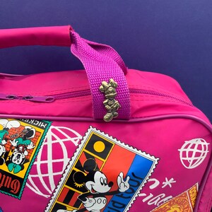 May include: Pink travel bag with a Mickey Mouse design. The bag has a zipper closure and a top handle. The handle has a small Minnie Mouse charm attached to it. The bag is decorated with colorful stamps featuring Mickey Mouse and the words "World Tour".