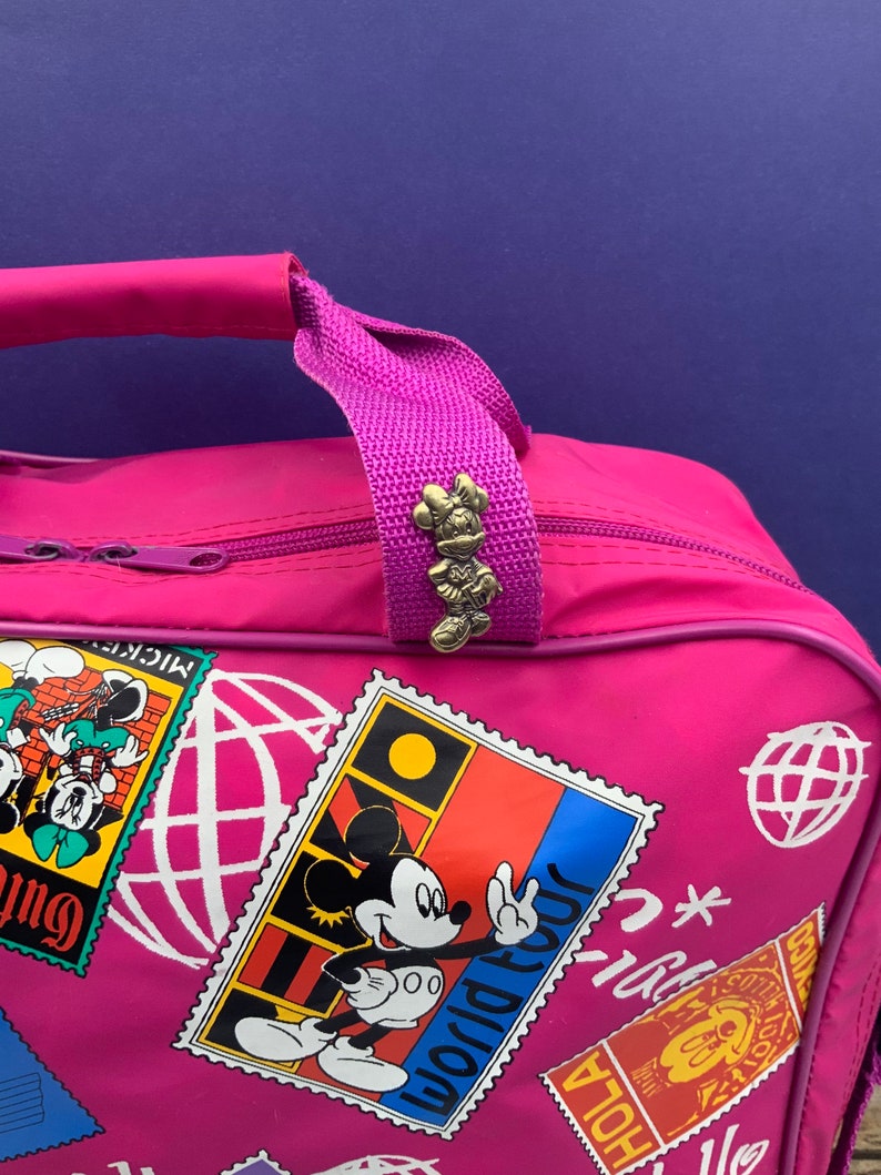 May include: Pink travel bag with a Mickey Mouse design. The bag has a zipper closure and a top handle. The handle has a small Minnie Mouse charm attached to it. The bag is decorated with colorful stamps featuring Mickey Mouse and the words "World Tour".