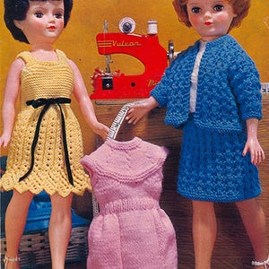 May include: Two dolls wearing knitted outfits. The doll on the left is wearing a yellow dress with a black sash. The doll on the right is wearing a blue cardigan and skirt. A sewing machine is in the background.
