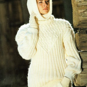 May include: A white knitted sweater with a turtleneck and a cable knit pattern. The sweater has a hood and is worn by a person with short brown hair.