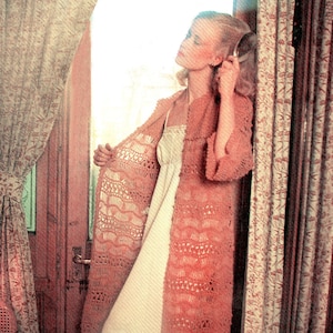 May include: A woman wearing a crocheted cardigan in a peachy pink colour. The cardigan has a delicate openwork pattern and is worn over a white slip dress.