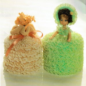 May include: Two crocheted tissue box covers, one in a light green colour with a doll wearing a green dress and hat, and the other in a cream colour with a bouquet of orange flowers.