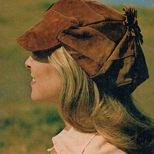 Puede incluir: Una mujer con el pelo largo y rubio lleva un sombrero de gamuza marrón con un gran lazo en un lado. El sombrero tiene una ala ancha y está ligeramente inclinado hacia adelante. La mujer mira hacia un lado, y su expresión es relajada.