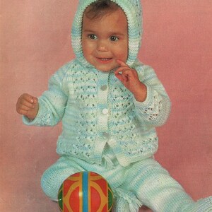 May include: A baby wearing a light blue and white striped knitted cardigan and matching trousers with a white button fastening. The baby is wearing a light blue knitted hat with a white trim. The baby is sitting on a pink surface and holding a red, yellow and blue ball.