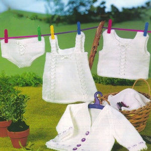 Puede incluir: Ropa de bebé de punto blanco colgada de un tendedero con pinzas. La ropa incluye un vestido sin mangas, un top sin mangas y una braga. Un cárdigan de punto blanco con botones morados está tirado en el suelo.