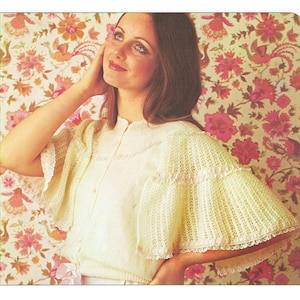 Puede incluir: Una capelina de punto amarillo claro con mangas acampanadas y cierre de botones en la parte delantera. La capelina presenta un delicado ribete de encaje y un diseño de punto suave y texturizado. El fondo es un papel pintado floral con flores rosas y melocotón.