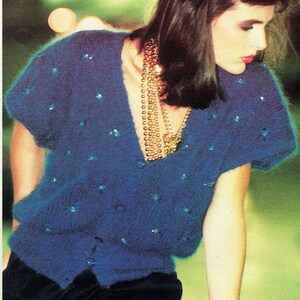 May include: A blue knit cardigan with short sleeves and sparkling embellishments. The cardigan has a V-neck and is buttoned down the front. A gold necklace and bracelet are also visible.