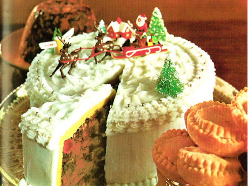 May include: A white frosted cake decorated with a miniature Santa Claus in a sleigh pulled by reindeer, a small green Christmas tree, and silver sprinkles. The cake is cut into slices and served with small, round, golden pastries.