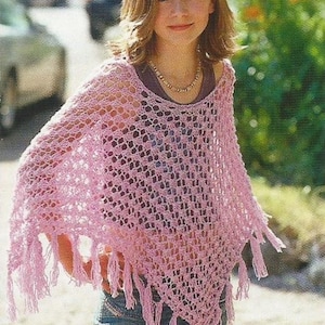 May include: A light pink, open-weave crochet poncho with fringe. The poncho is worn over a dark top and blue jeans. A necklace is visible. The overall style is casual and bohemian.