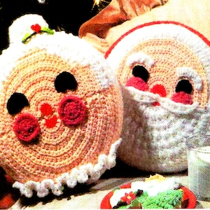 May include: Two crocheted Christmas cushions, one shaped like a gingerbread person and the other like Santa Claus. The gingerbread person cushion is pink and white with black eyes and red cheeks. The Santa Claus cushion is white with a red hat and a white beard.