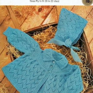 Puede incluir: Un conjunto de punto azul para bebé compuesto por un cárdigan y un gorro con un patrón de rombos. El conjunto está colocado sobre un lecho de paja en una cesta de madera. El número de patrón es 1999 y el precio es de 10 peniques.