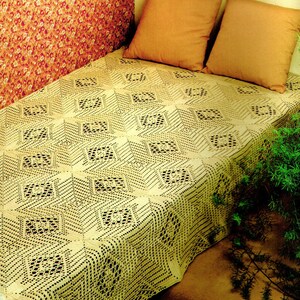 Op de afbeelding: Een gehaakt deken met een kant-vierkant patroon in een crème kleur. Het deken ligt op een bed met twee oranje kussens en de tekst "Lacy Squares" staat onderaan de afbeelding.