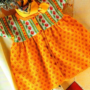 Puede incluir: Vestido de bebé tejido en tonos amarillos y naranjas, adornado con un estampado floral. El corpiño presenta un diseño a rayas con flores rojas, rayas verdes y amarillas. Se incluye un gorro a juego con un lazo.