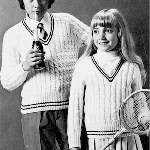 May include: Two children wearing white cable knit sweaters with black and red trim. The boy is holding a bottle of soda and a straw. The girl is holding a tennis racket.