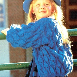 May include: A young girl wearing a blue knitted cardigan with a cable knit pattern and a blue hat. She is wearing a black and white striped skirt and is holding onto a green railing.