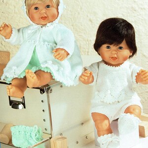 Könnte beinhalten: Zwei Babypuppen in handgestrickten Outfits. Eine Puppe trägt einen hellblauen Kapuzenmantel und Shorts, die andere einen weißen Strampler und Socken. Die Puppen sitzen auf einer weißen Oberfläche mit Holzblöcken.