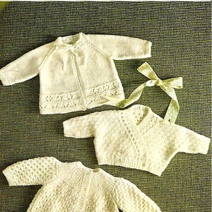 May include: Three white knitted baby sweaters with different stitch patterns. The sweaters are laid out on a green textured surface. The text "Shepherd 62 Double Knitting" is visible in the top right corner of the image.
