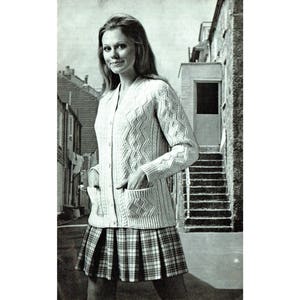 May include: A white cable knit cardigan with a v-neck and pockets, worn over a plaid skirt.